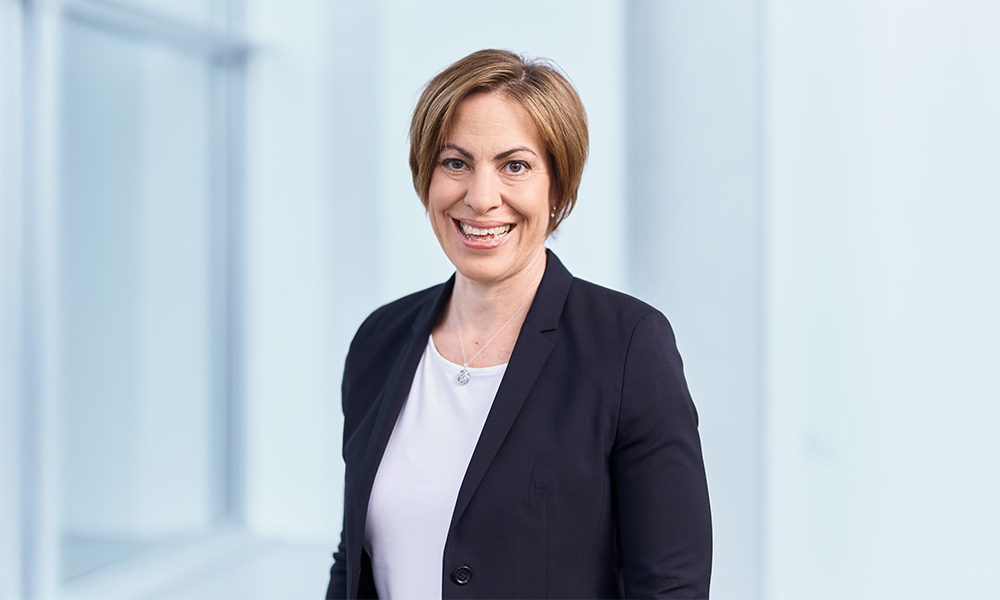 Smiling woman in a dark blazer and white top standing in a bright office corridor.