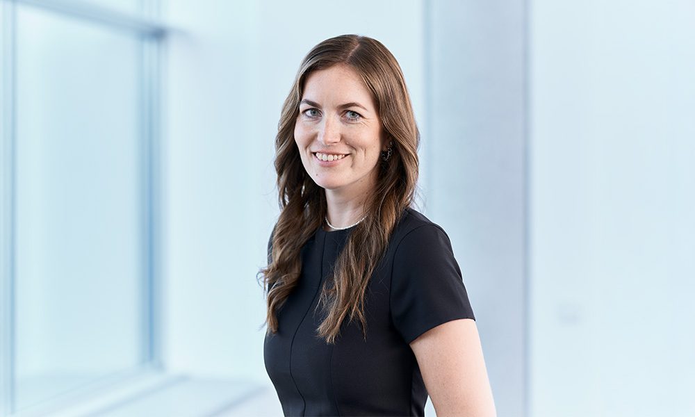 Portrait of a smiling woman in a dark professional dress standing in a bright office area, facing slightly to the side.
