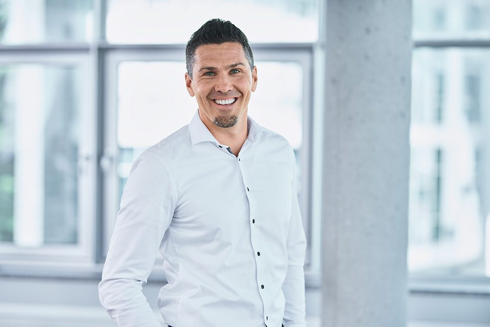 Smiling man in a white button-down shirt standing in a bright office with large windows behind him.