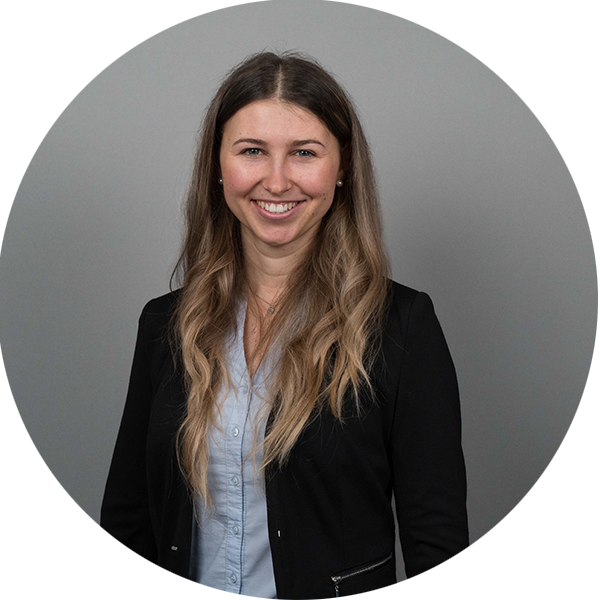 lisa-kuebler Portrait of a smiling woman with long wavy hair, wearing a light blue blouse and black blazer against a gray backdrop.